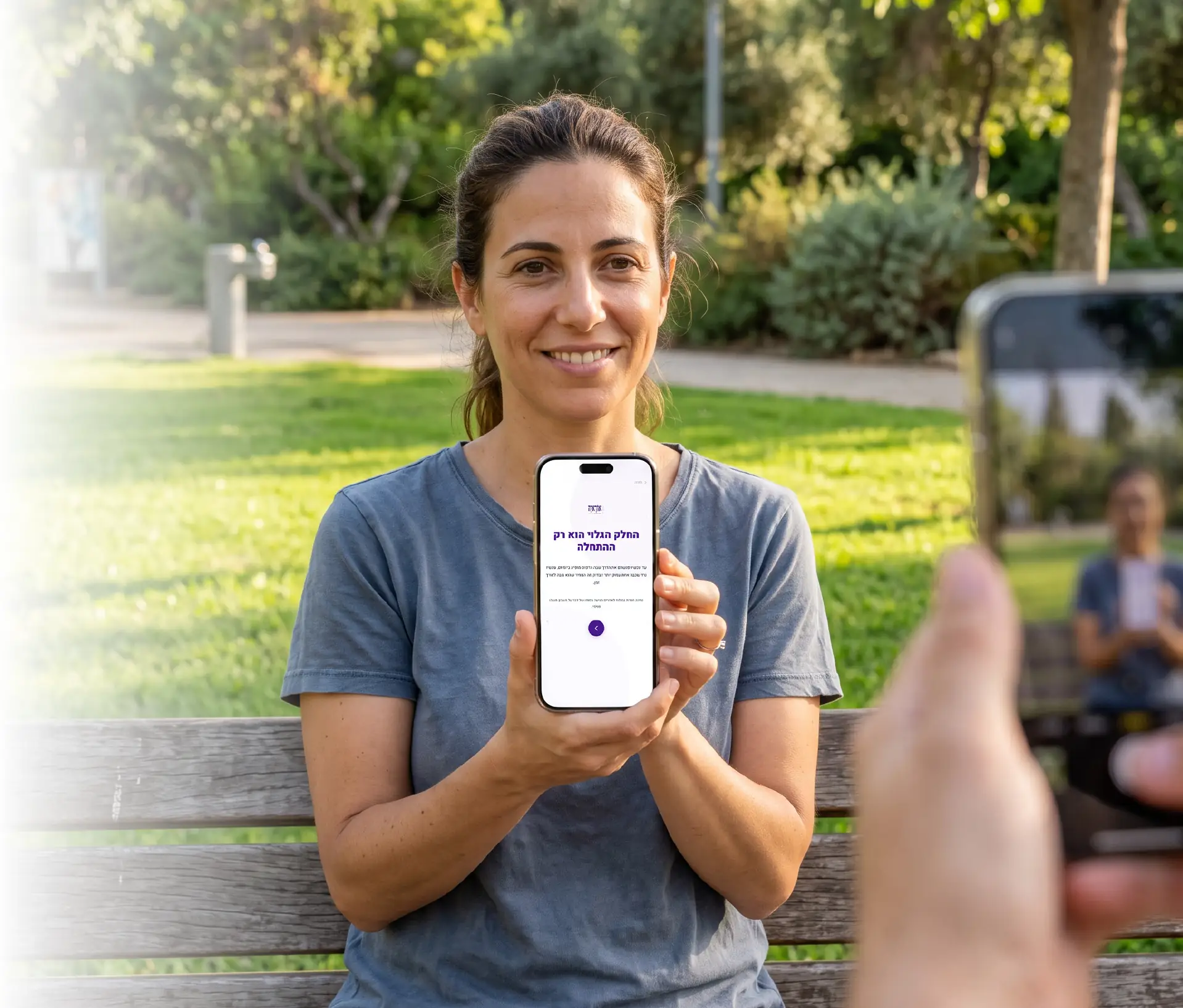 אישה יושבת על ספסל בפארק ומראה את השאלון במסך הטלפון